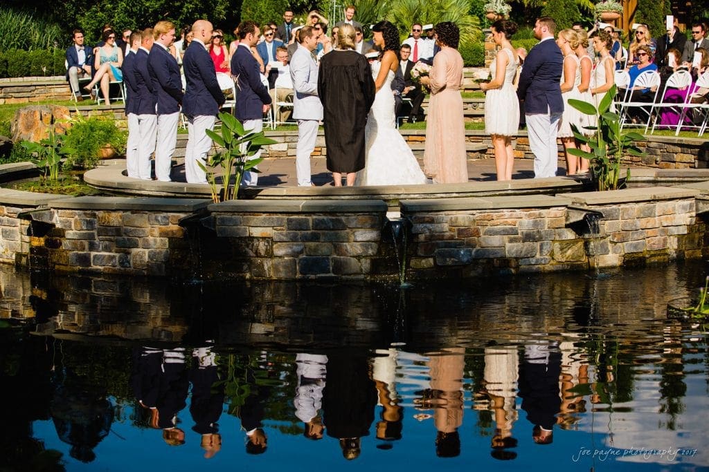 duke gardens wedding photography - jordan & zeke A wedding ceremony outdoors on a sunny day, featuring a couple exchanging vows on a circular platform surrounded by water. The bridal party, dressed in formal wear, stands beside them. Guests seated in the background. Reflection visible in the water.
