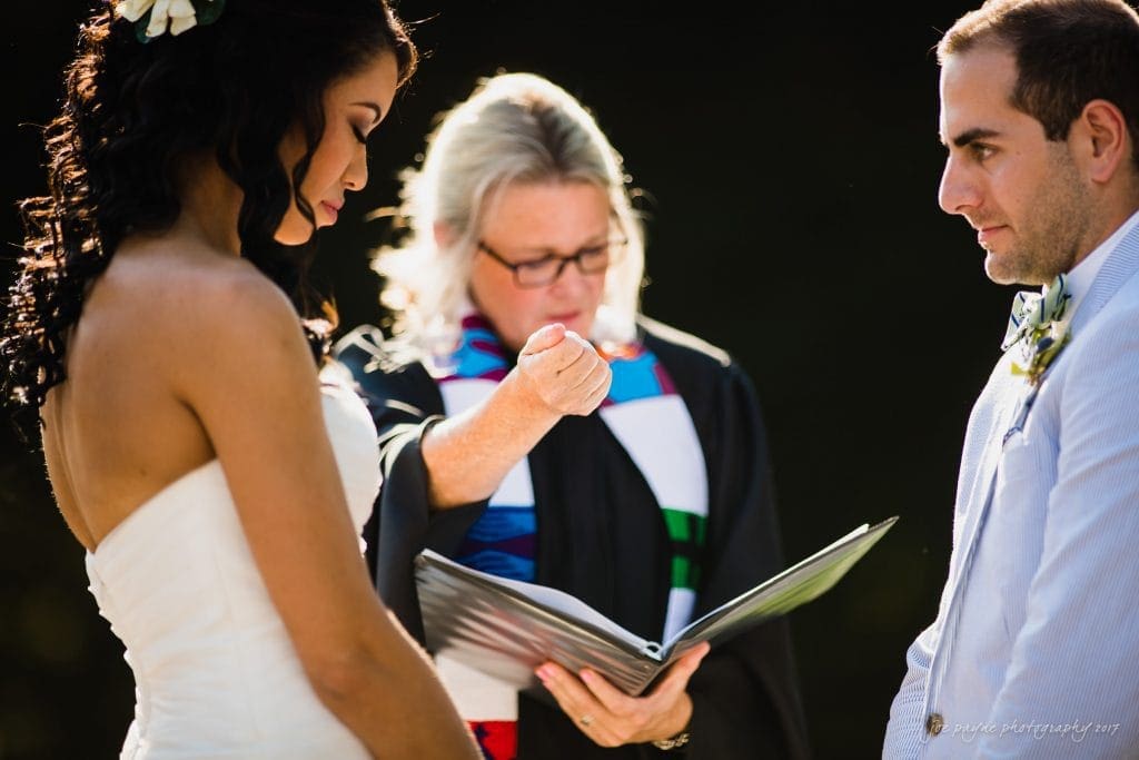 duke gardens wedding photography - jordan & zeke A couple stands facing each other during a wedding ceremony. The officiant, in a robe with a colorful stole, gestures while holding an open book. The bride wears a strapless gown, and the groom is in a light suit. There are flowers in the brides hair.