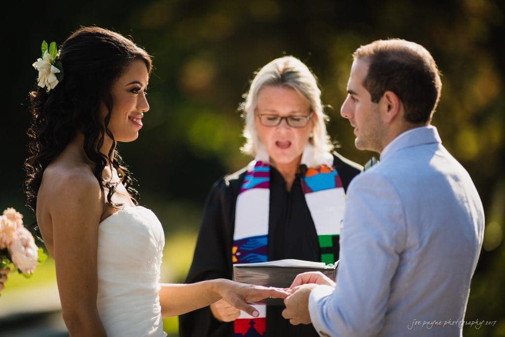 duke gardens wedding photography - jordan & zeke A bride and groom exchange rings during an outdoor wedding ceremony. The officiant stands between them, wearing glasses and a colorful stole. The bride holds a small bouquet and wears a white dress with a flower in her hair. The groom wears a light suit.