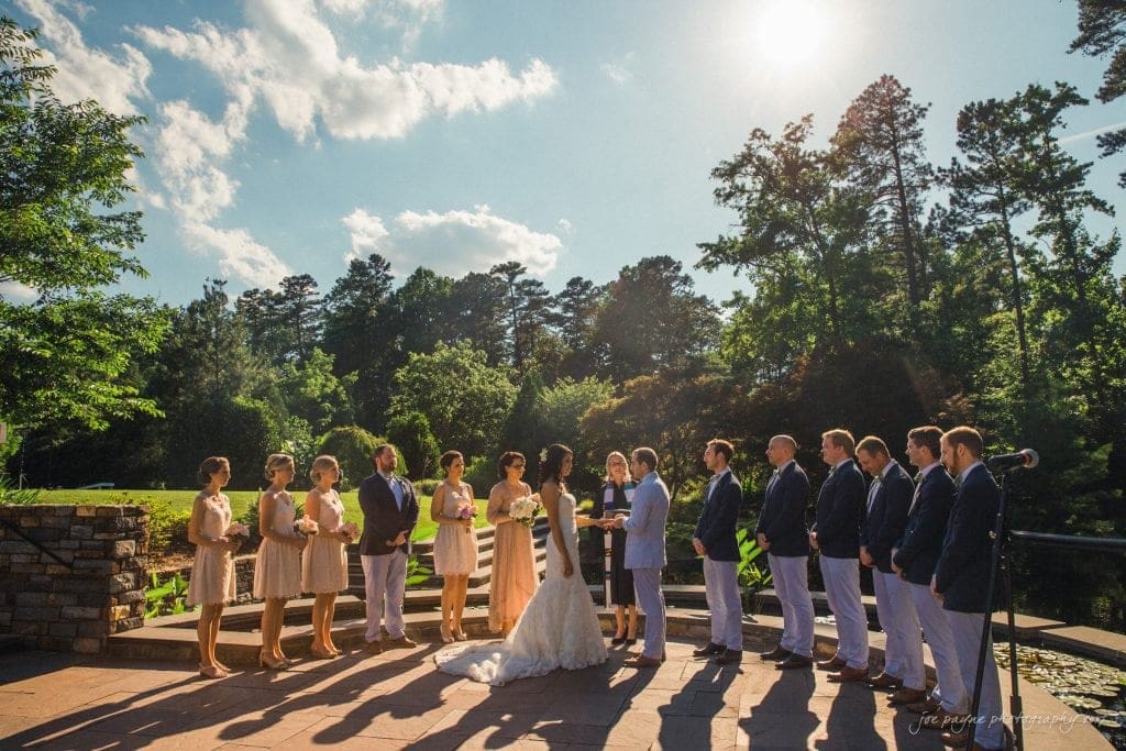 duke gardens wedding photography - jordan & zeke A wedding ceremony is taking place outdoors on a sunny day. The bride and groom stand in the center, surrounded by bridesmaids and groomsmen. The lush greenery and clear sky create a picturesque backdrop.