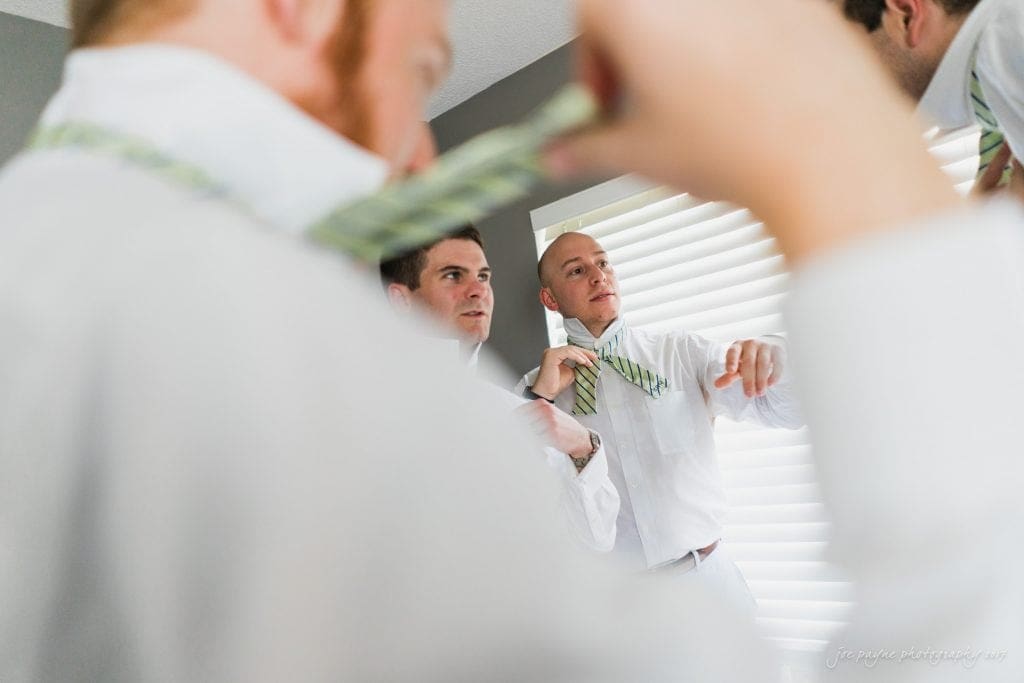 duke gardens wedding photography - jordan & zeke A group of men in white shirts and green-striped ties are gathering in a bright room, with one adjusting his tie. The focus is on their preparation and conversation, with sunlight filtering through a window in the background.