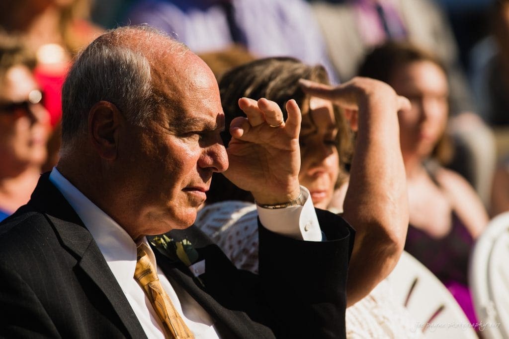 duke gardens wedding photography - jordan & zeke An elderly man in a suit and tie and a woman shielding their eyes from the sun with their hands. They are seated outdoors at an event, surrounded by other people. The sunlight creates shadows on their faces.