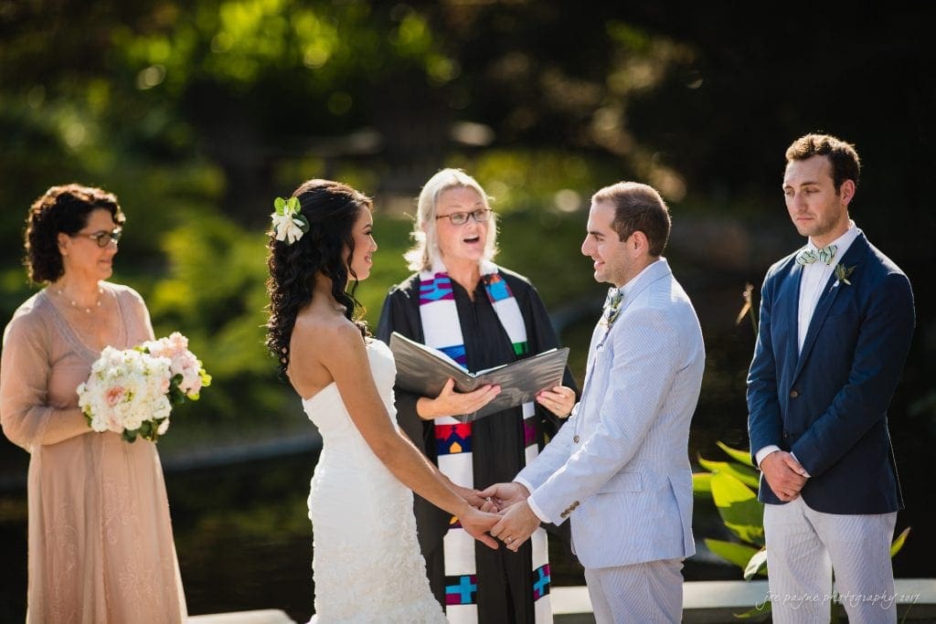 duke gardens wedding photography - jordan & zeke A couple holds hands during a wedding ceremony. The bride wears a white dress and the groom a light suit, with a pond in the background. An officiant and two attendants stand nearby, one holding a bouquet.
