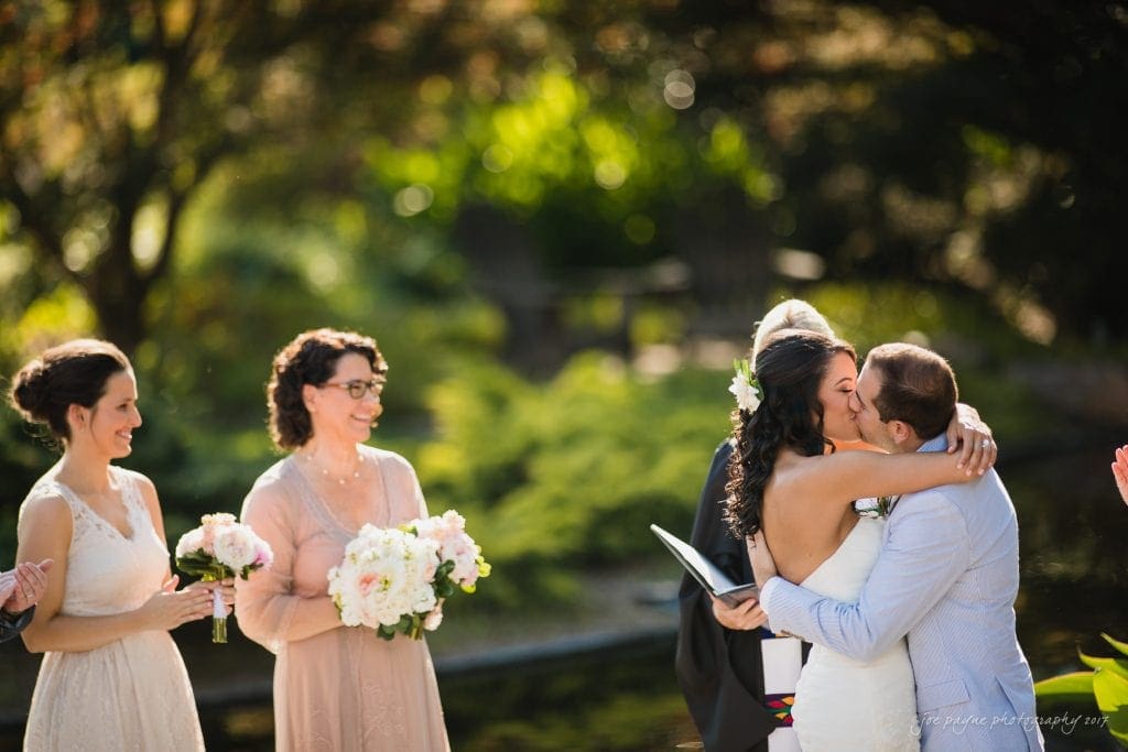 duke gardens wedding photography - jordan & zeke A couple shares a kiss during an outdoor wedding ceremony. The bride, in a white dress, holds a bouquet and embraces the groom, dressed in a light suit. Bridesmaids and a guest stand nearby, smiling with bouquets. Lush greenery surrounds them.