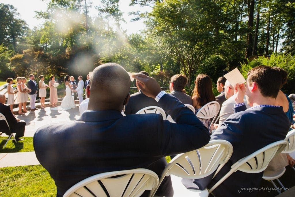 duke gardens wedding photography - jordan & zeke Outdoor wedding ceremony with guests seated in white chairs. A man in a suit shades his eyes while watching the couple and wedding party at the altar. Sunlight filters through trees, creating a warm, inviting atmosphere.