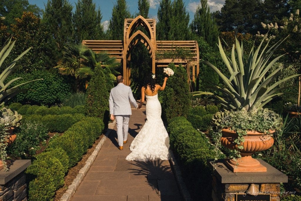 duke gardens wedding photography - jordan & zeke A newlywed couple joyfully walks hand in hand down a garden path. The bride holds her bouquet aloft, and they move toward a wooden trellis surrounded by lush greenery and potted plants under a clear blue sky.