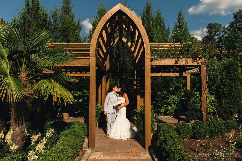 duke gardens wedding photography - jordan & zeke A bride in a white dress and groom in a light gray suit share a kiss under a wooden pergola in a lush garden. Sunlight illuminates the scene, highlighting the greenery and ornate surroundings.