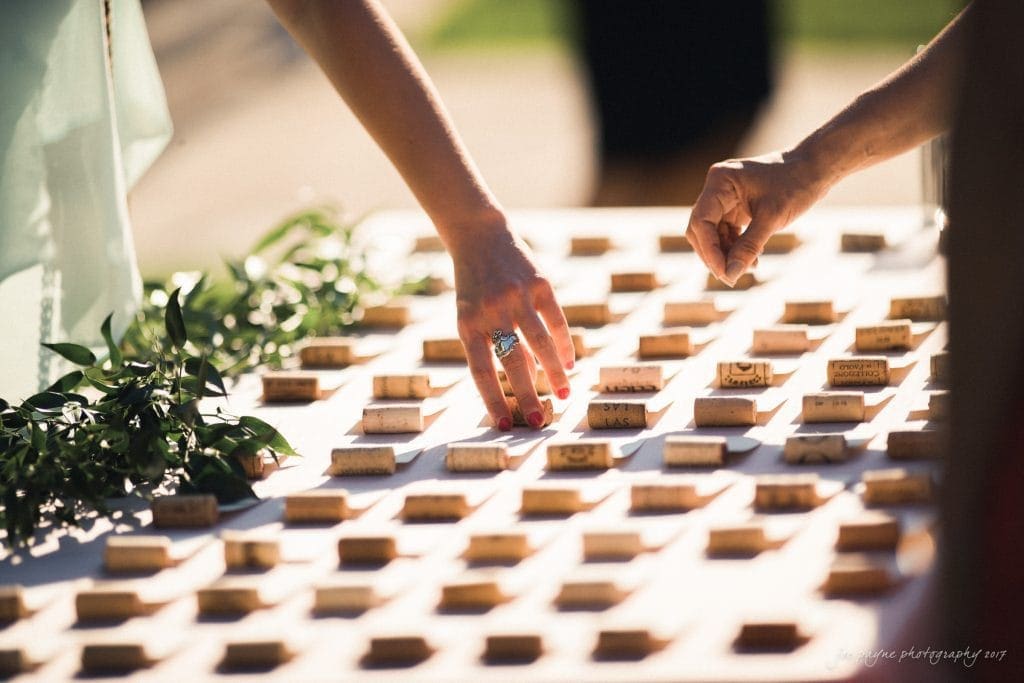 Two hands reach for corks on a table arranged in a grid pattern. One hand wears a ring and red nail polish. Greenery is placed on the tables left side, and warm sunlight casts soft shadows.