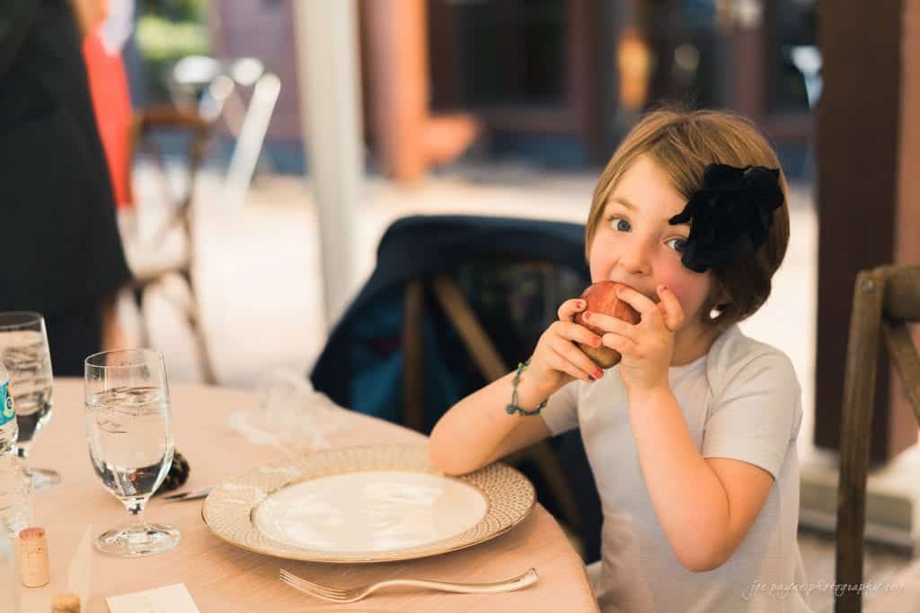 duke gardens wedding photography - jordan & zeke A young girl with short hair and a black flower accessory is sitting at a dining table. She is smiling and holding a bread roll close to her face. The table is set with a plate and glassware. The background is softly blurred.