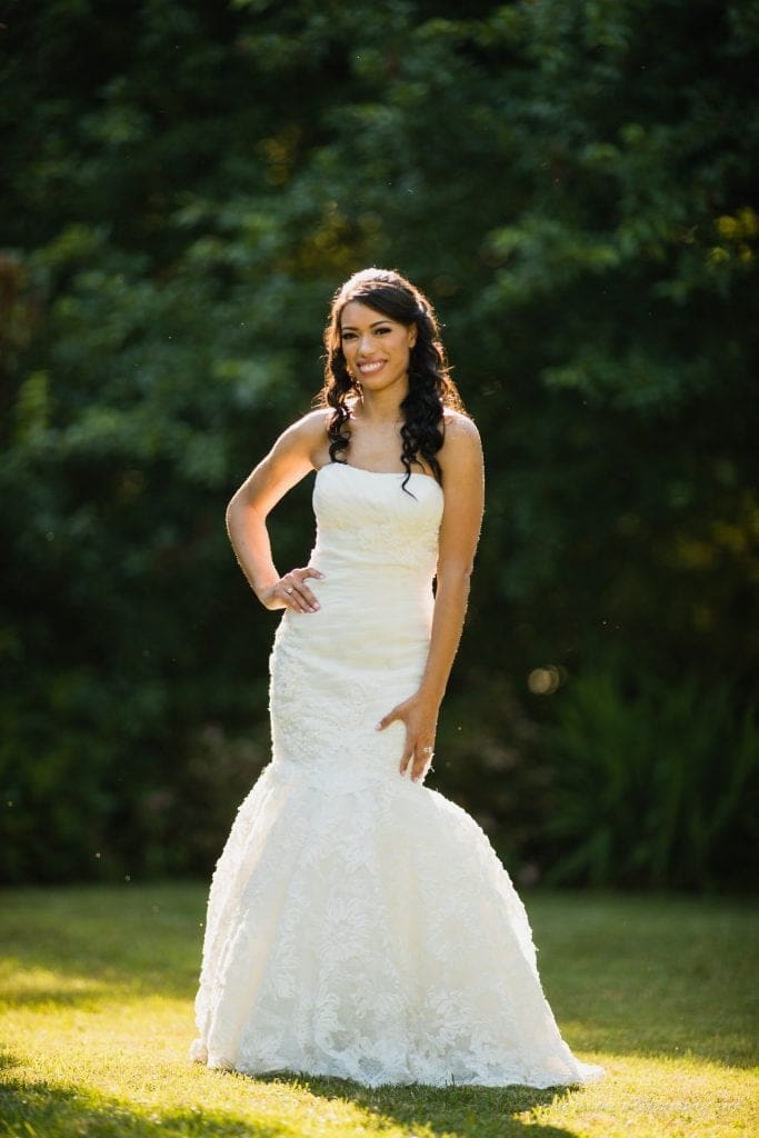 duke gardens wedding photography - jordan & zeke A bride in a lace wedding gown poses outdoors on a sunny day. She stands on grass with lush green trees in the background, smiling and resting her hand on her hip. Her long hair is styled with gentle curls.