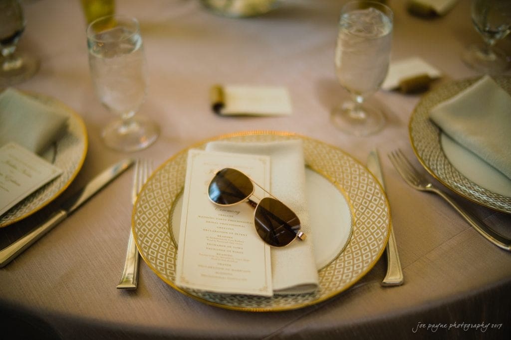 duke gardens wedding photography - jordan & zeke A pair of sunglasses rests on a folded napkin atop a dinner menu on an elegant table setting. The table features fine china with a geometric pattern, cutlery, and glasses filled with water. The ambiance is warm and sophisticated.