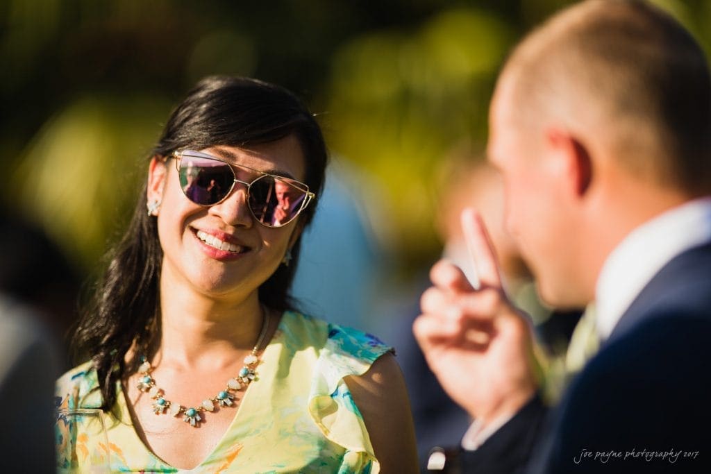 duke gardens wedding photography - jordan & zeke A woman wearing sunglasses and a floral dress smiles outdoors, engaged in a conversation with a man in a suit. The sunlight creates a warm ambiance, and the background is softly blurred.