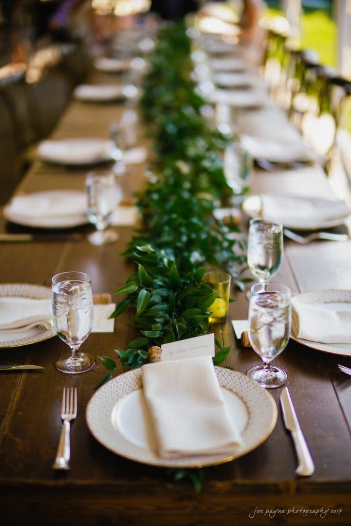 A long wooden dining table is elegantly set with white plate settings and napkins. Glasses of water are placed beside each setting. A lush green foliage runner adorns the center of the table, creating a sophisticated and inviting atmosphere.