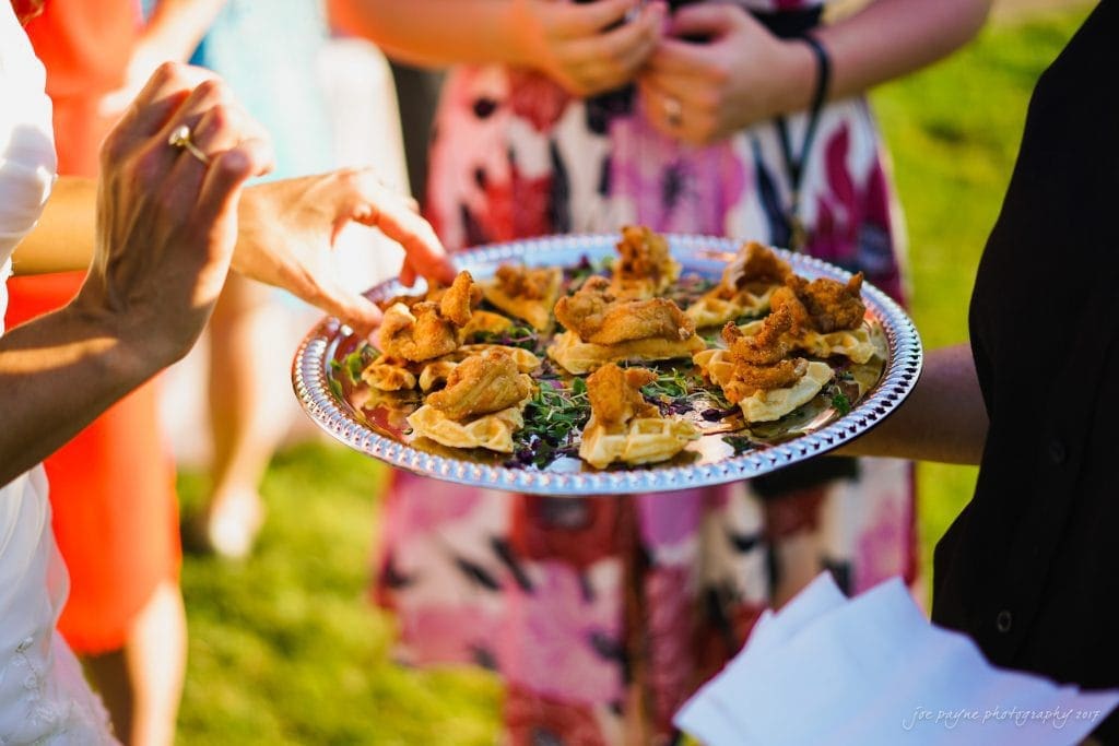 duke gardens wedding photography - jordan & zeke A person holds a silver tray of savory appetizers outdoors. The appetizers are arranged neatly and appear to contain fried items on a base. Several people stand nearby in colorful clothing. The grass is visible in the background.