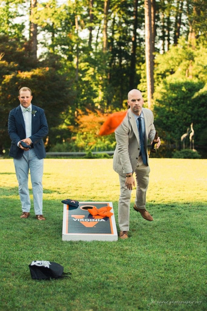 duke gardens wedding photography - jordan & zeke Two men play cornhole on a grassy lawn. One throws an orange beanbag towards a board with Virginia written on it. Both are dressed in suits, with trees and greenery in the background.