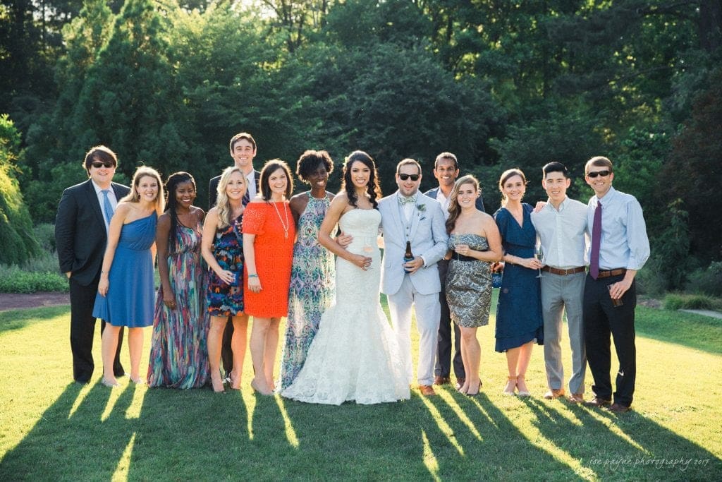A group of people dressed in formal attire, including a bride in a white gown, pose together outdoors on a sunny day. They stand on a grassy area with trees in the background. Everyone is smiling or looking at the camera.