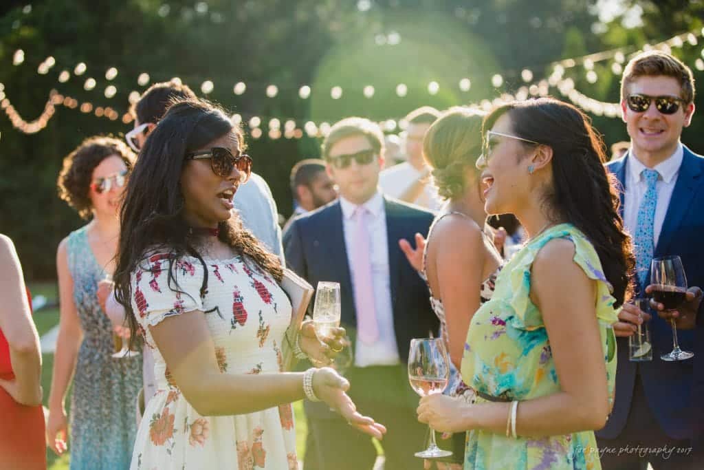 duke gardens wedding photography - jordan & zeke A group of people dressed in formal attire are enjoying an outdoor event. Two women in dresses are conversing and holding wine glasses, while others are mingling in the background. String lights are visible above them.