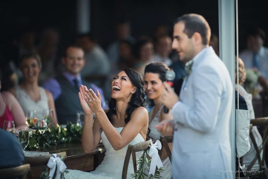 duke gardens wedding photography - jordan & zeke A bride claps and smiles joyfully while seated at a wedding reception table. Guests around her also appear happy and engaged. She is focused on a man in a suit who is giving a speech with a microphone. The atmosphere is festive and lively.