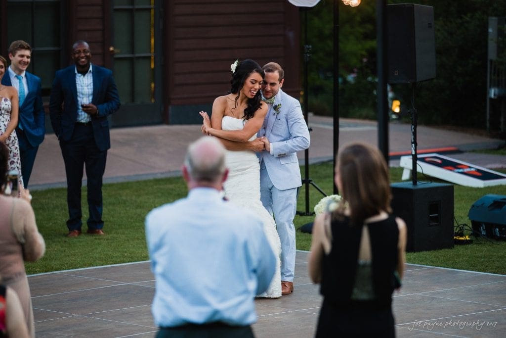 duke gardens wedding photography - jordan & zeke A bride and groom share their first dance outdoors on a wooden floor, surrounded by guests. The bride wears a white gown, and the groom is in a light-colored suit. Onlookers watch and take photos, with a building and greenery in the background.
