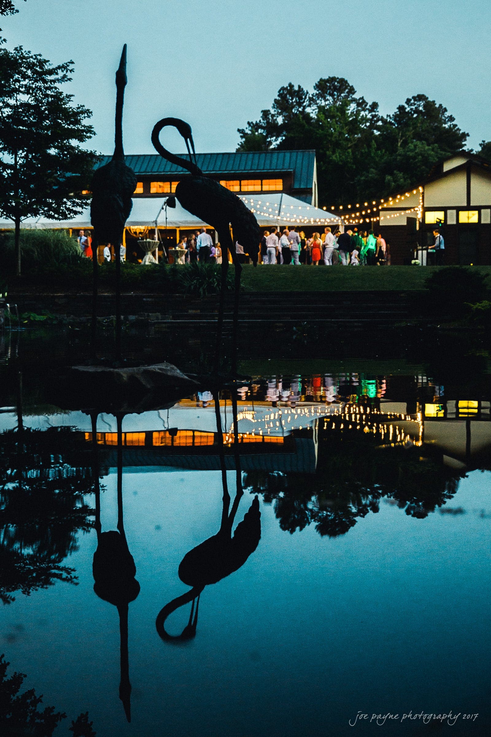 duke gardens wedding photography - jordan & zeke Silhouettes of bird sculptures reflected in a pond at dusk, with a warmly lit pavilion and string lights in the background, hosting a gathering of people. Trees and sky add to the serene atmosphere.