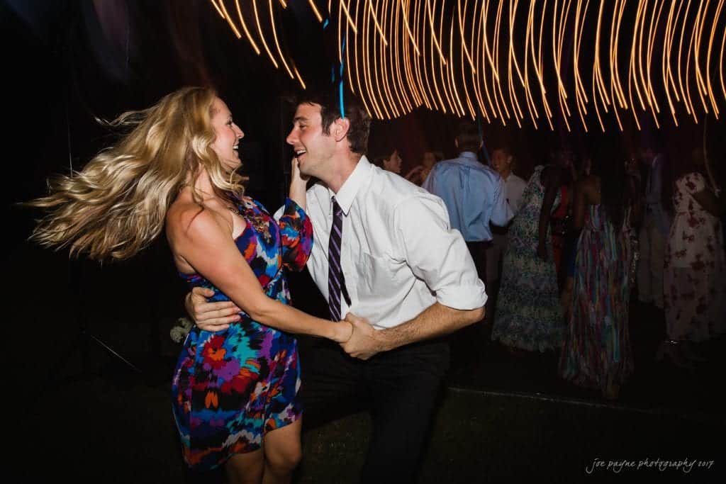 duke gardens wedding photography - jordan & zeke A woman in a colorful dress dances energetically with a man in a white shirt and tie at an outdoor event. The background is blurred with streaks of light, creating a lively atmosphere.