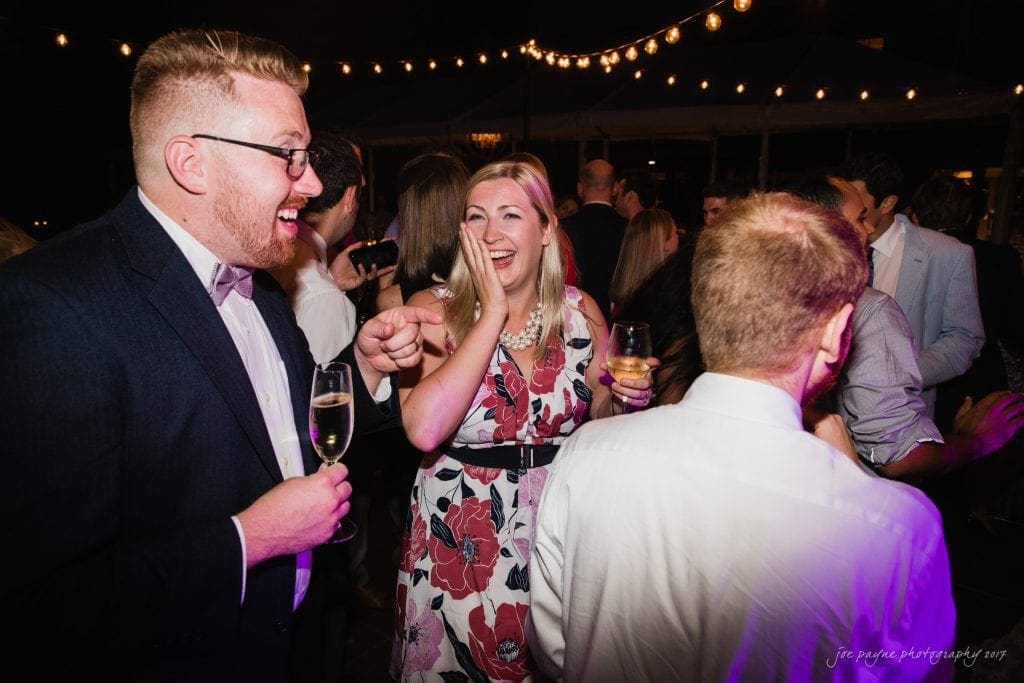 duke gardens wedding photography - jordan & zeke A group of people at a lively outdoor night event. A man in a suit and a woman in a floral dress are laughing and holding drinks. String lights hang above, creating a warm and festive atmosphere.