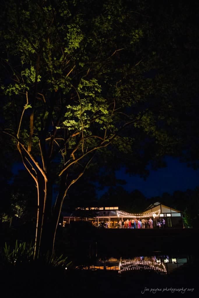 duke gardens wedding photography - jordan & zeke A nighttime scene shows a tree in the foreground and a building in the background, lit by string lights. People are gathered inside the building, and their reflections are visible in a body of water below.