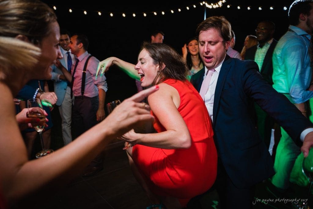 duke gardens wedding photography - jordan & zeke A lively dance floor scene at a party, featuring a woman in a red dress energetically dancing next to a man in a suit. People in the background also appear to be enjoying themselves, with festive lights overhead creating a joyful atmosphere.