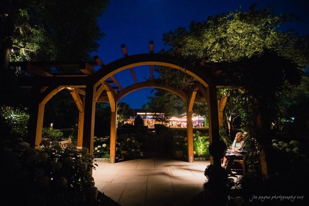 duke gardens wedding photography - jordan & zeke A woman sits on a bench under a wooden pergola at night. The background is illuminated with soft lights from a distant building, surrounded by trees and lush greenery, creating a serene and peaceful atmosphere.