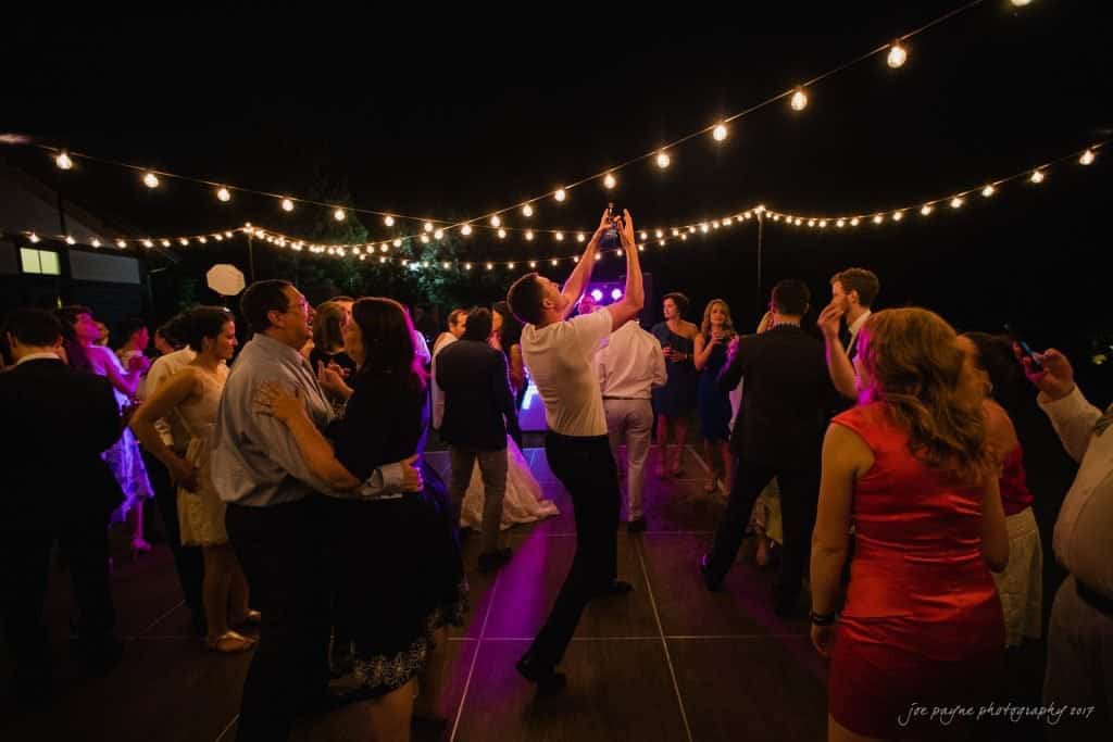 duke gardens wedding photography - jordan & zeke A lively outdoor party at night with people dancing under string lights. The central figure takes a photo with a smartphone. Attendees are dressed in formal and semi-formal outfits, creating a festive atmosphere.