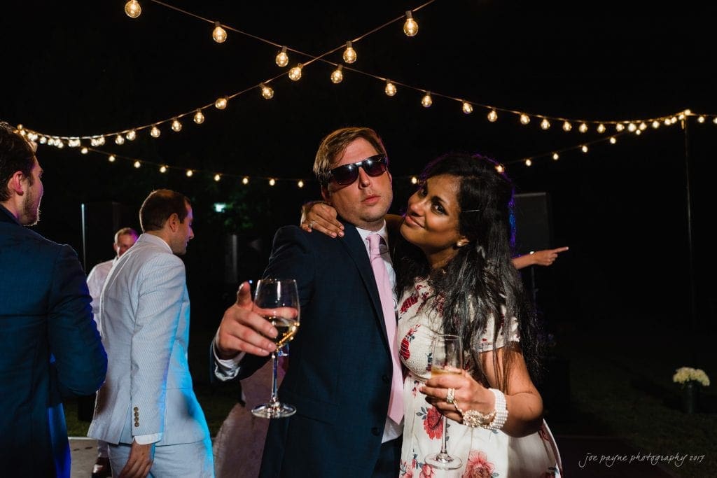 duke gardens wedding photography - jordan & zeke A couple poses playfully in a dimly lit outdoor setting with string lights overhead. The man wears sunglasses and points at the camera while holding a wine glass. The woman, dressed in a floral outfit, leans on him smiling, also holding a glass.