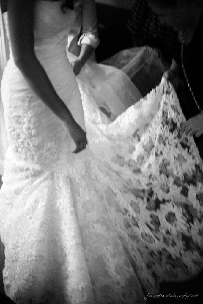 duke gardens wedding photography - jordan & zeke A black and white photo of a bride wearing a lace wedding gown. Two people assist her, adjusting the train of the dress. The fabric features intricate lace patterns, adding elegance to the scene.