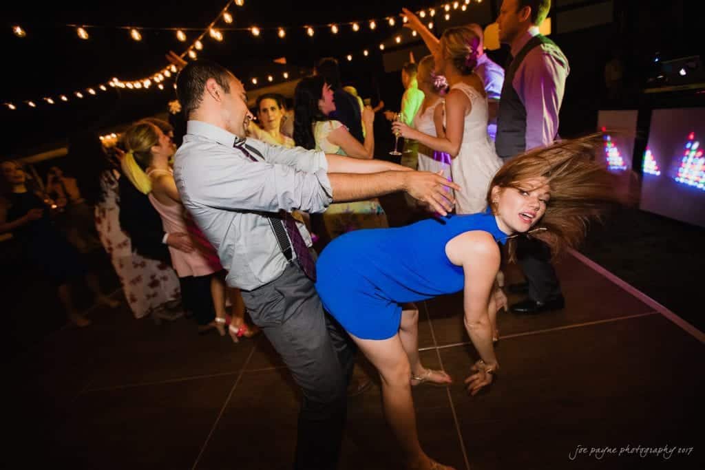 duke gardens wedding photography - jordan & zeke A lively wedding reception with people energetically dancing under string lights. A man and a woman in a blue dress are dancing closely, surrounded by other guests dressed in formal attire. DJ booth with colorful lights is visible in the background.