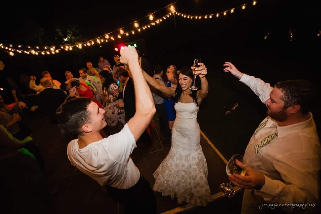 duke gardens wedding photography - jordan & zeke A joyful bride in a white dress and groom are dancing energetically under string lights at an outdoor wedding reception. Guests surround them, clapping and dancing, with drinks in hand, creating a festive atmosphere on a wooden dance floor.