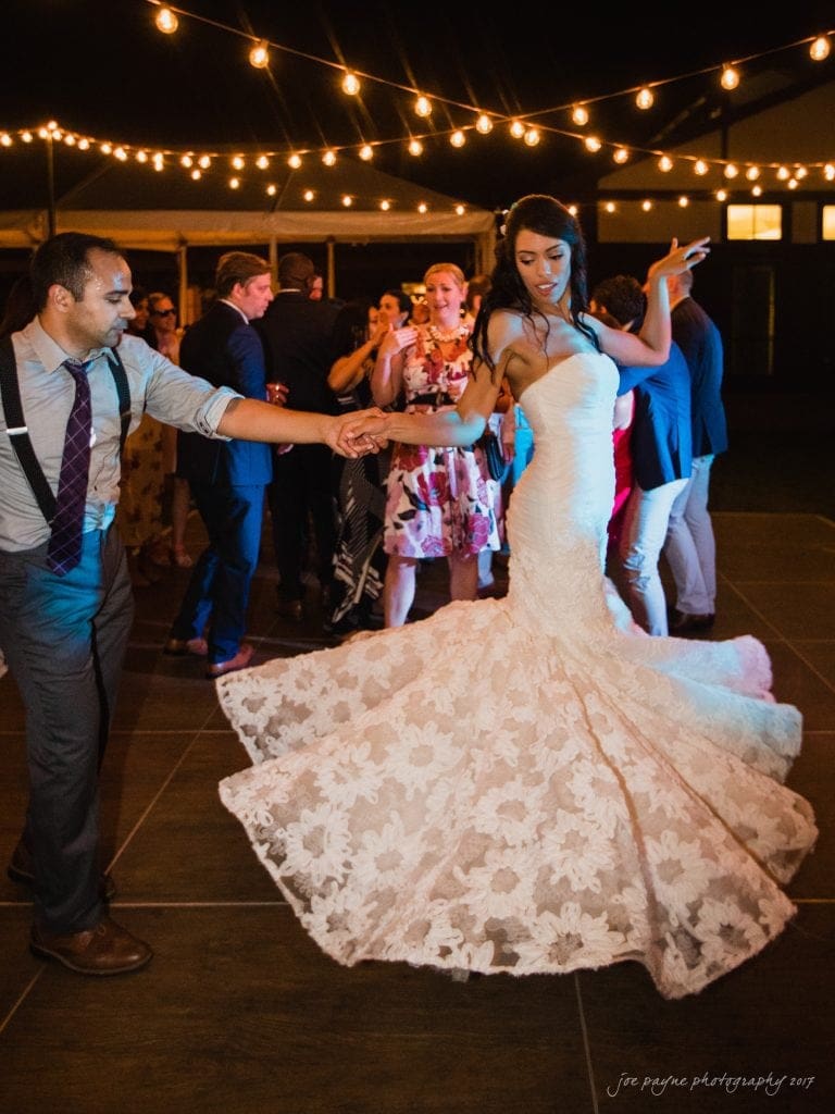 duke gardens wedding photography - jordan & zeke A bride in a flowing lace gown dances with a man in suspenders on an outdoor dance floor. String lights illuminate the night scene, and guests are gathered in the background enjoying the celebration.