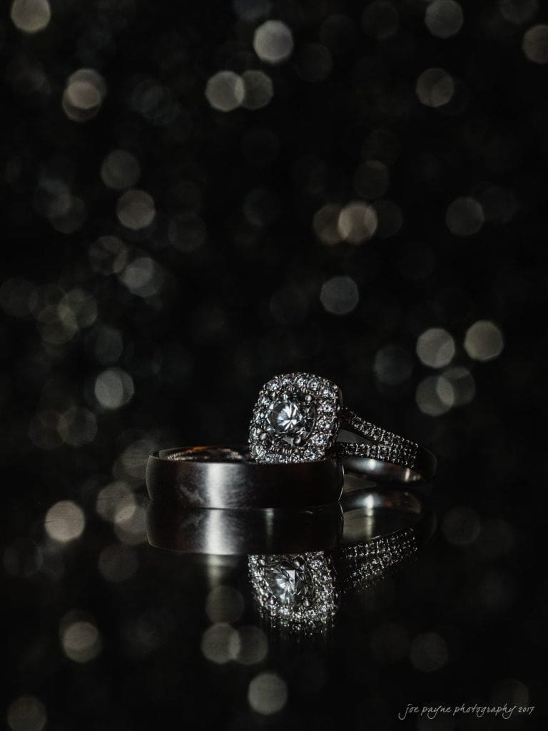 duke gardens wedding photography - jordan & zeke A pair of elegant rings resting on a reflective surface. One ring is a simple band, and the other features a dazzling diamond with smaller stones around it. The background is dark with soft-focus lights creating a bokeh effect.