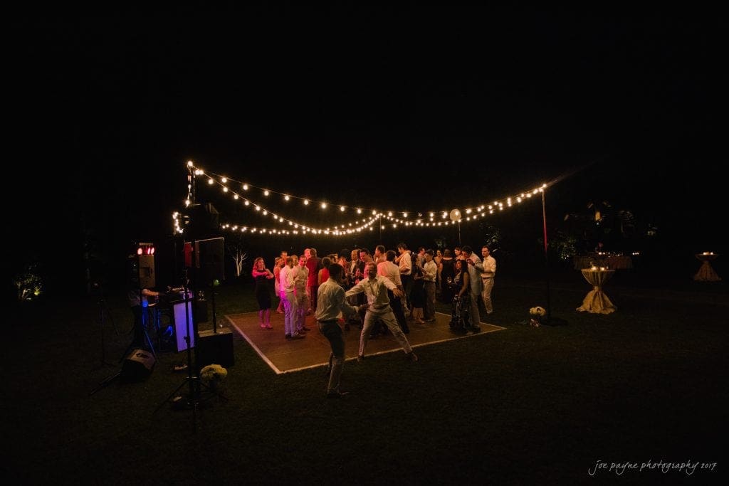 duke gardens wedding photography - jordan & zeke A group of people dancing under string lights on a dark outdoor dance floor. The setting is festive, with warm lighting creating a lively atmosphere. A DJ setup is visible in the corner, and trees can be seen in the background.