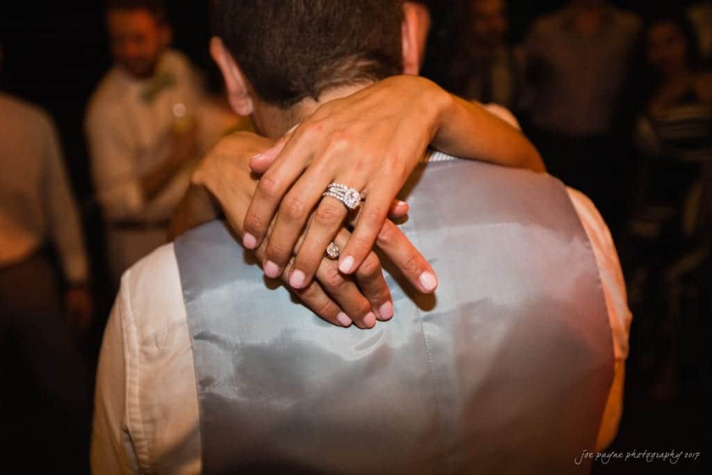 duke gardens wedding photography - jordan & zeke A close-up of a couple dancing, with the woman’s hands wrapped around the mans neck. Her fingers have a wedding and engagement ring. The man is wearing a light gray vest. People in the background are slightly blurred.