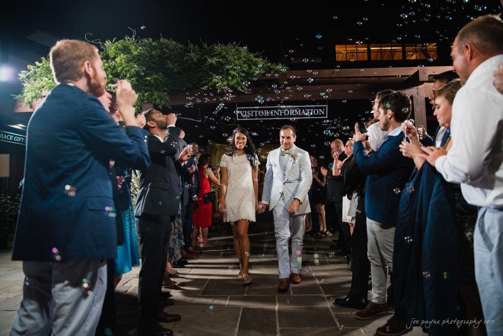duke gardens wedding photography - jordan & zeke A couple walks hand in hand through a joyful crowd blowing bubbles at night. The bride wears a short white dress, and the groom wears a light gray suit. The scene is festive with smiles and bubbly ambiance. A Visitor Information sign is in the background.