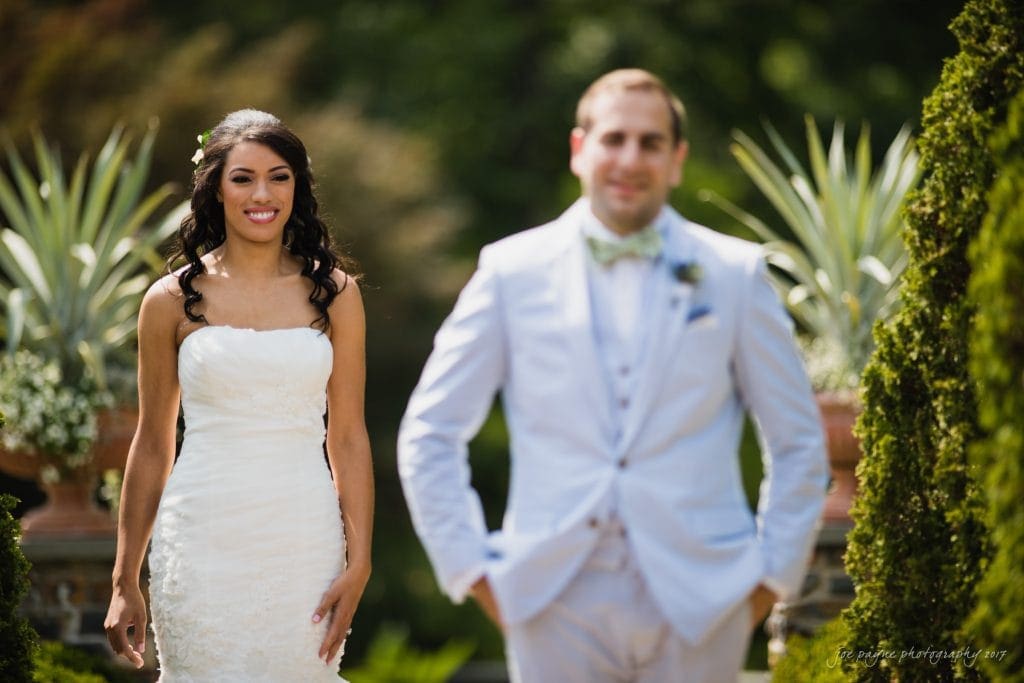 duke gardens wedding photography - jordan & zeke A smiling bride in a white dress stands outdoors, with greenery in the background. A man in a light-colored suit is in the foreground, slightly out of focus. The setting appears to be a garden.