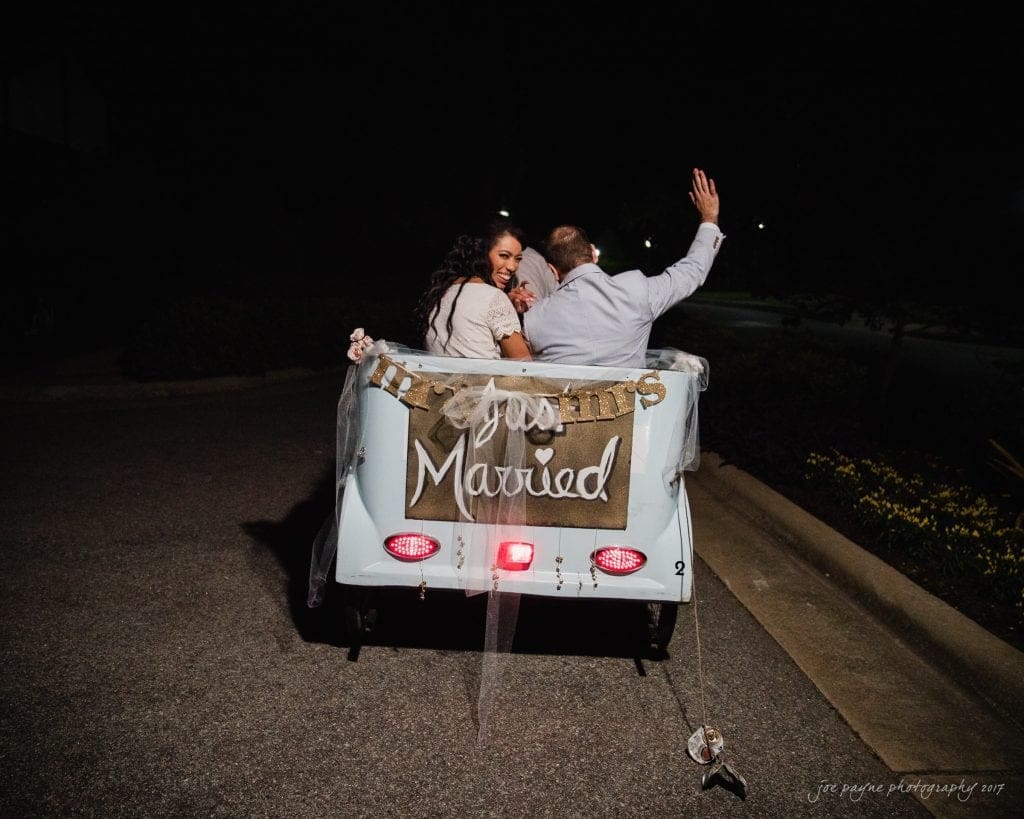 duke gardens wedding photography - jordan & zeke A newlywed couple in a small blue car waves as they drive away. The car features a Just Married sign on the back. Its nighttime, and the taillights illuminate the scene. The couple appears joyful and celebratory.