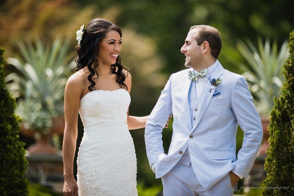 duke gardens wedding photography - jordan & zeke A bride in a white strapless gown and a groom in a light gray suit with a bow tie stand outside, smiling and looking at each other. The background is lush with greenery, creating a serene and joyful atmosphere.
