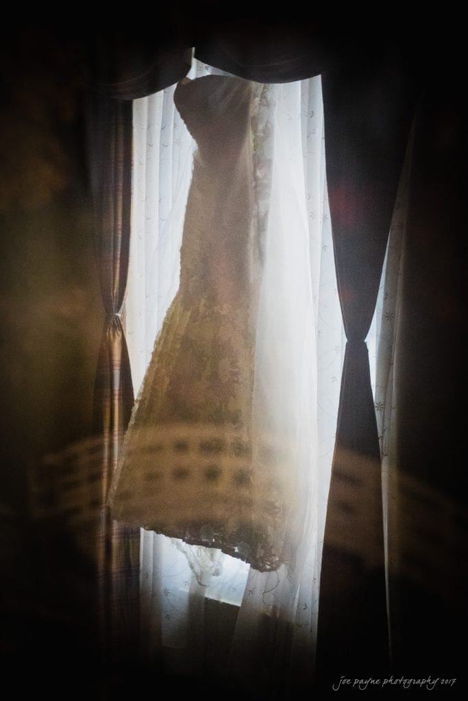 duke gardens wedding photography - jordan & zeke A wedding dress is elegantly silhouetted against a softly lit window, with sheer curtains creating a delicate backdrop. The dress features intricate lace details, highlighted by the gentle glow of the light behind.