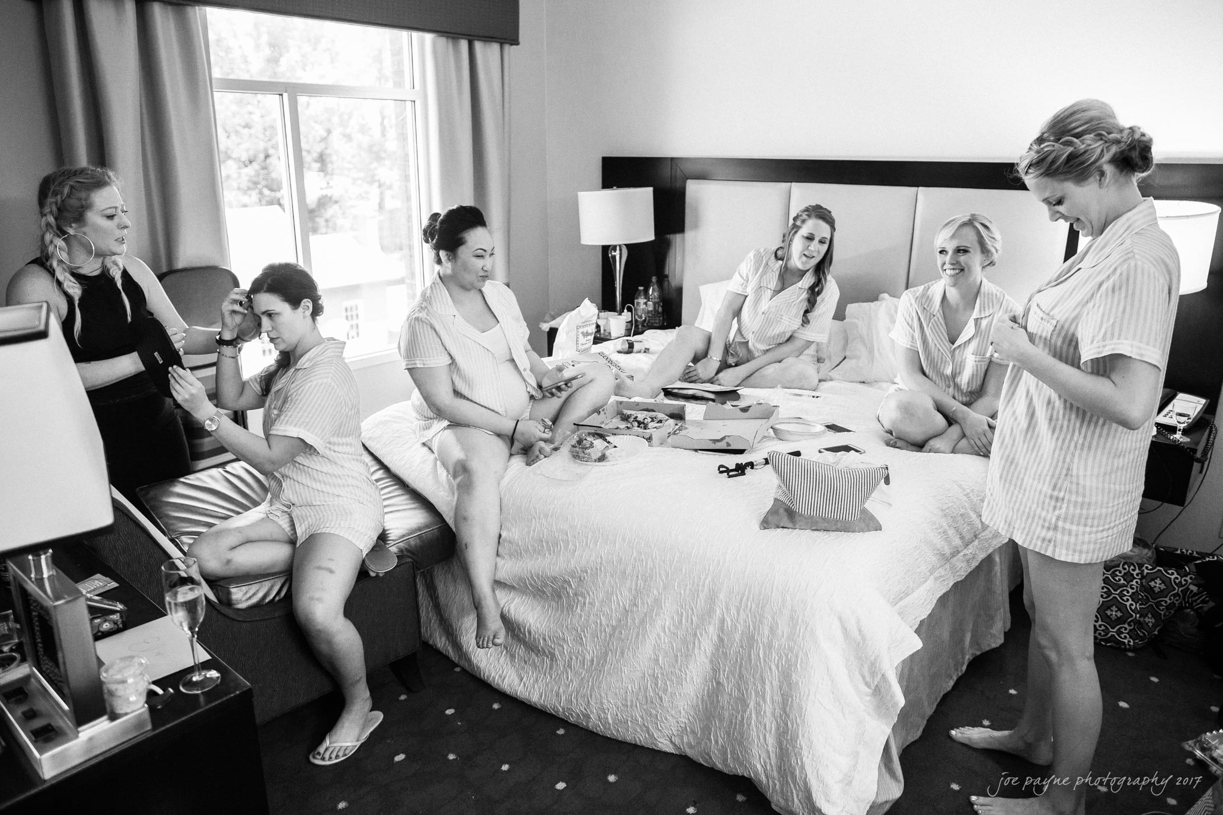 melrose knitting mill wedding - natalie & elliot A group of six women in casual clothing are in a hotel room. They are gathered around a bed, some sitting, others standing. They appear to be preparing for an event, with makeup and hair styling in progress. The room is well-lit with natural light.