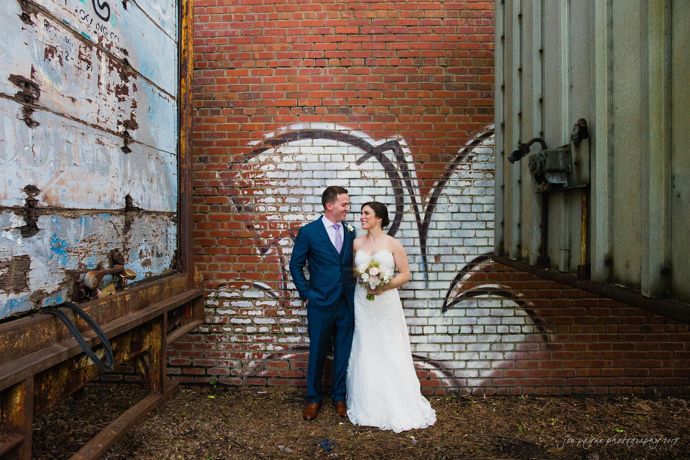 melrose knitting mill wedding - natalie & elliot A bride and groom stand smiling next to each other against a brick wall with graffiti. The bride holds a bouquet of flowers and wears a white dress, while the groom is in a blue suit with a red tie. The setting appears urban and artistic.