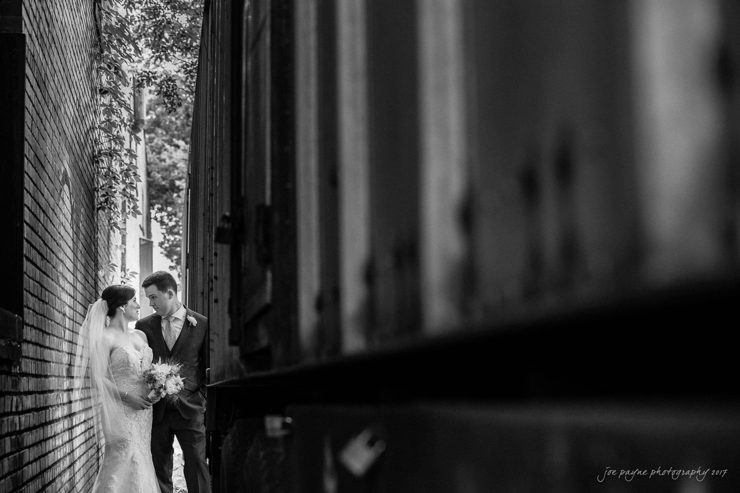 melrose knitting mill wedding - natalie & elliot A black and white photo of a bride and groom standing closely together in an alleyway, surrounded by brick walls and a train car. The bride is in a wedding dress holding a bouquet, and the groom is in a suit. Trees can be seen in the background.