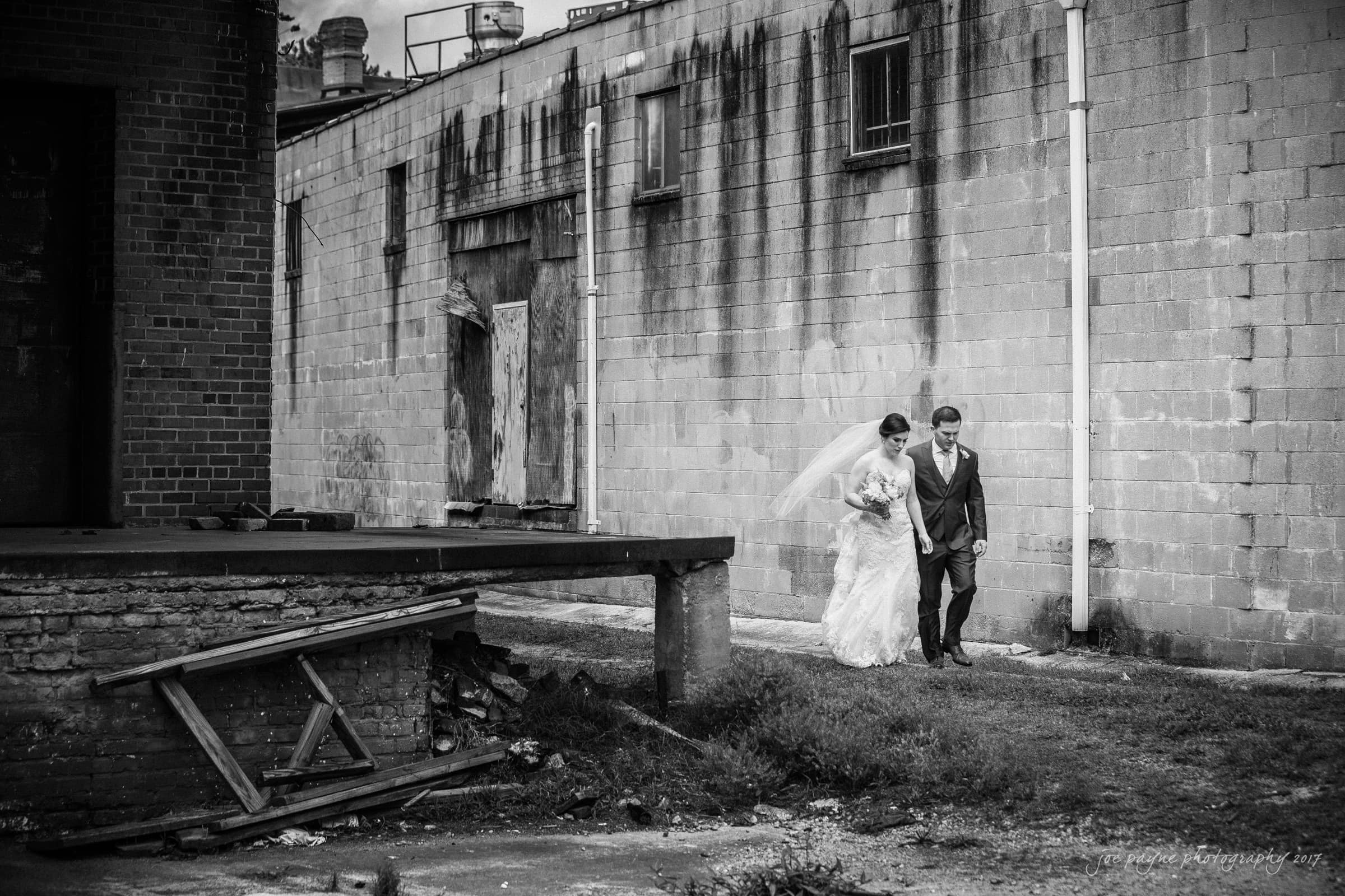 melrose knitting mill wedding - natalie & elliot A bride and groom walk hand in hand along a worn, industrial building. The bride wears a veil and holds flowers. The groom is in a suit and tie. The scene is in black and white, giving it a timeless feel.