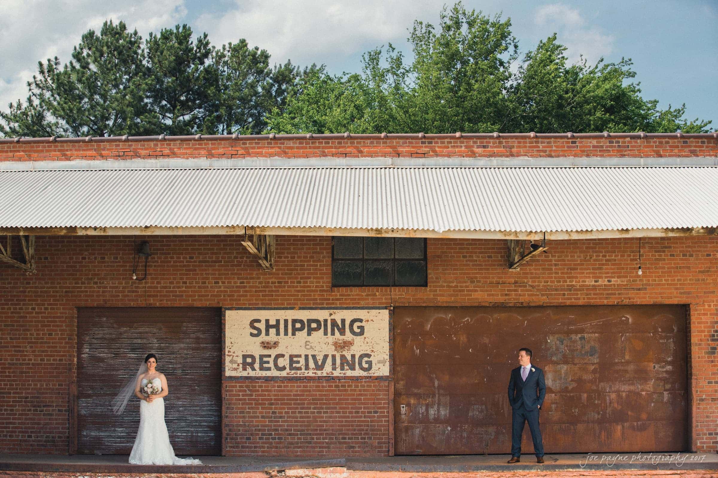 melrose knitting mill wedding - natalie & elliot A bride in a white dress and a groom in a suit stand apart outside a large brick building. The building has a sign that reads Shipping Receiving. Trees are visible in the background under a clear blue sky.