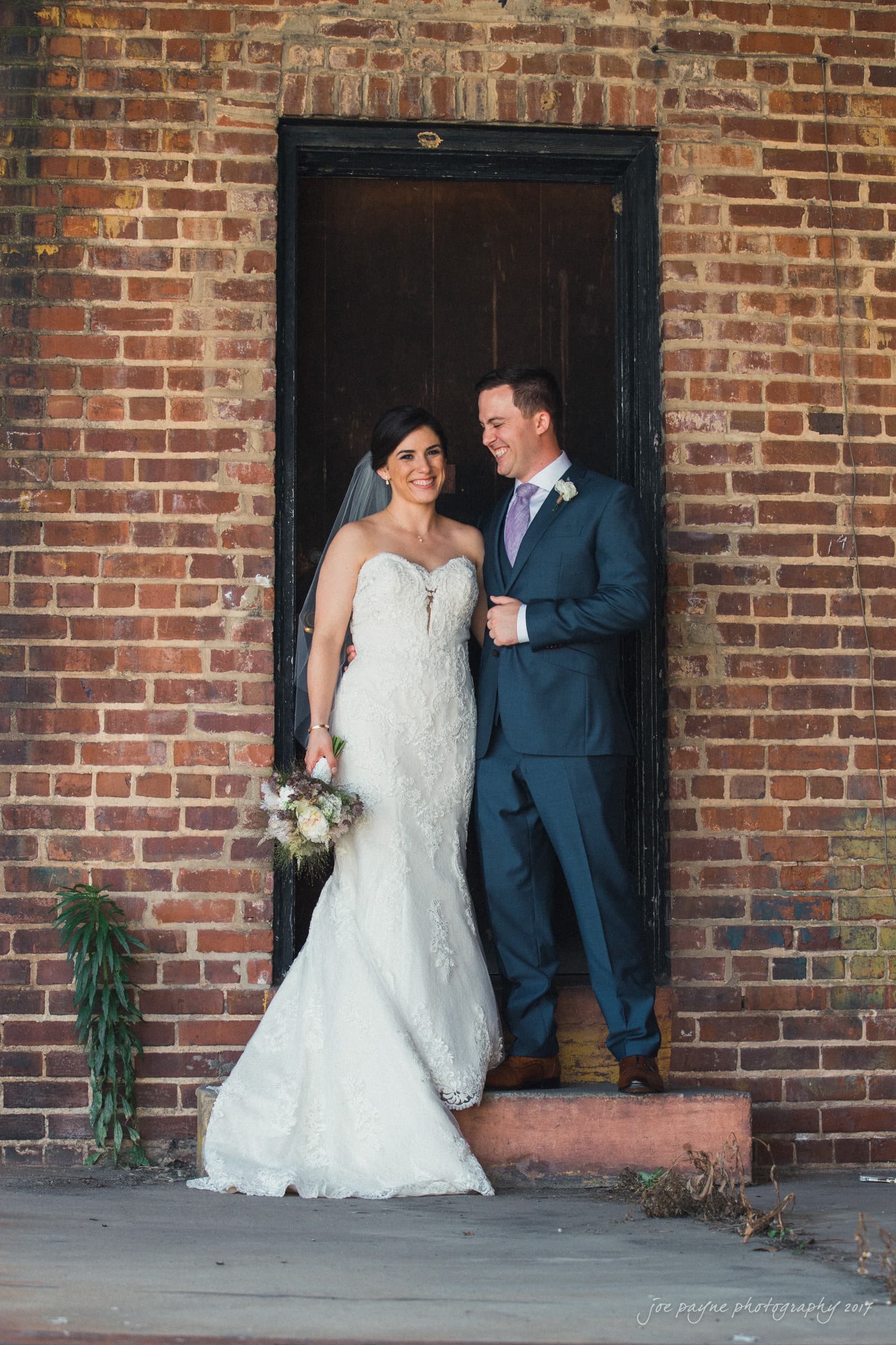 melrose knitting mill wedding - natalie & elliot A bride in a lace gown holds a bouquet while standing with a groom in a navy suit and purple tie. They are smiling and standing in a brick doorway.