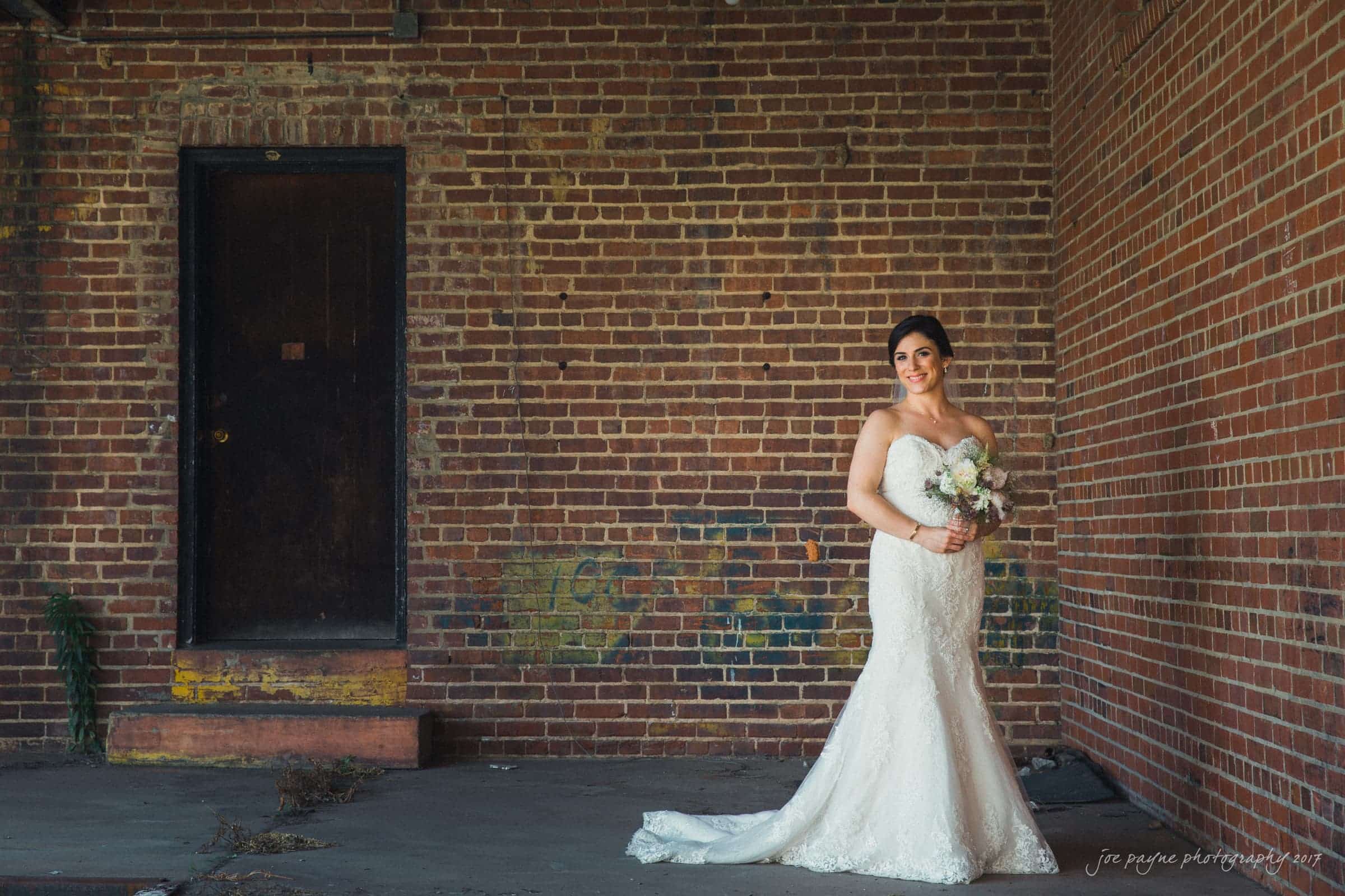 melrose knitting mill wedding - natalie & elliot A bride in a white gown stands against a rustic, brick wall. She holds a bouquet and smiles, creating a contrast between her elegant attire and the urban, industrial setting.